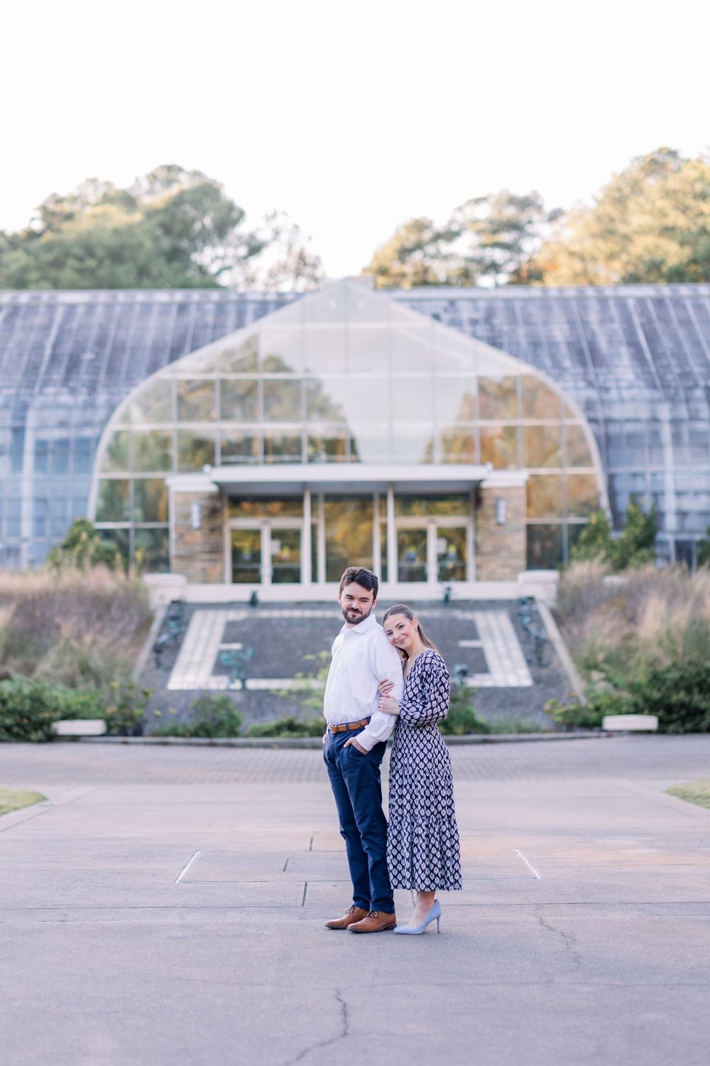 A couple poses for engagement photos in front of a large greenhouse structure at sunset.