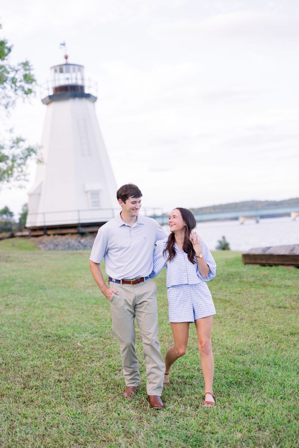A couple in coordinated white and blue outfits walks together on a grassy hill with a lighthouse in the background.
