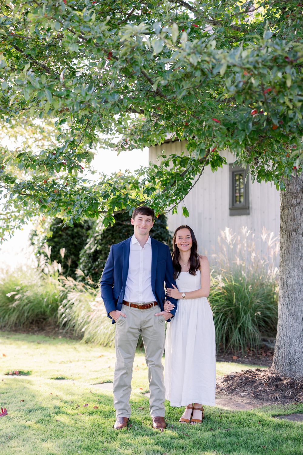 A couple stands together in an outdoor garden setting surrounded by lush green trees and grass on a sunny day.