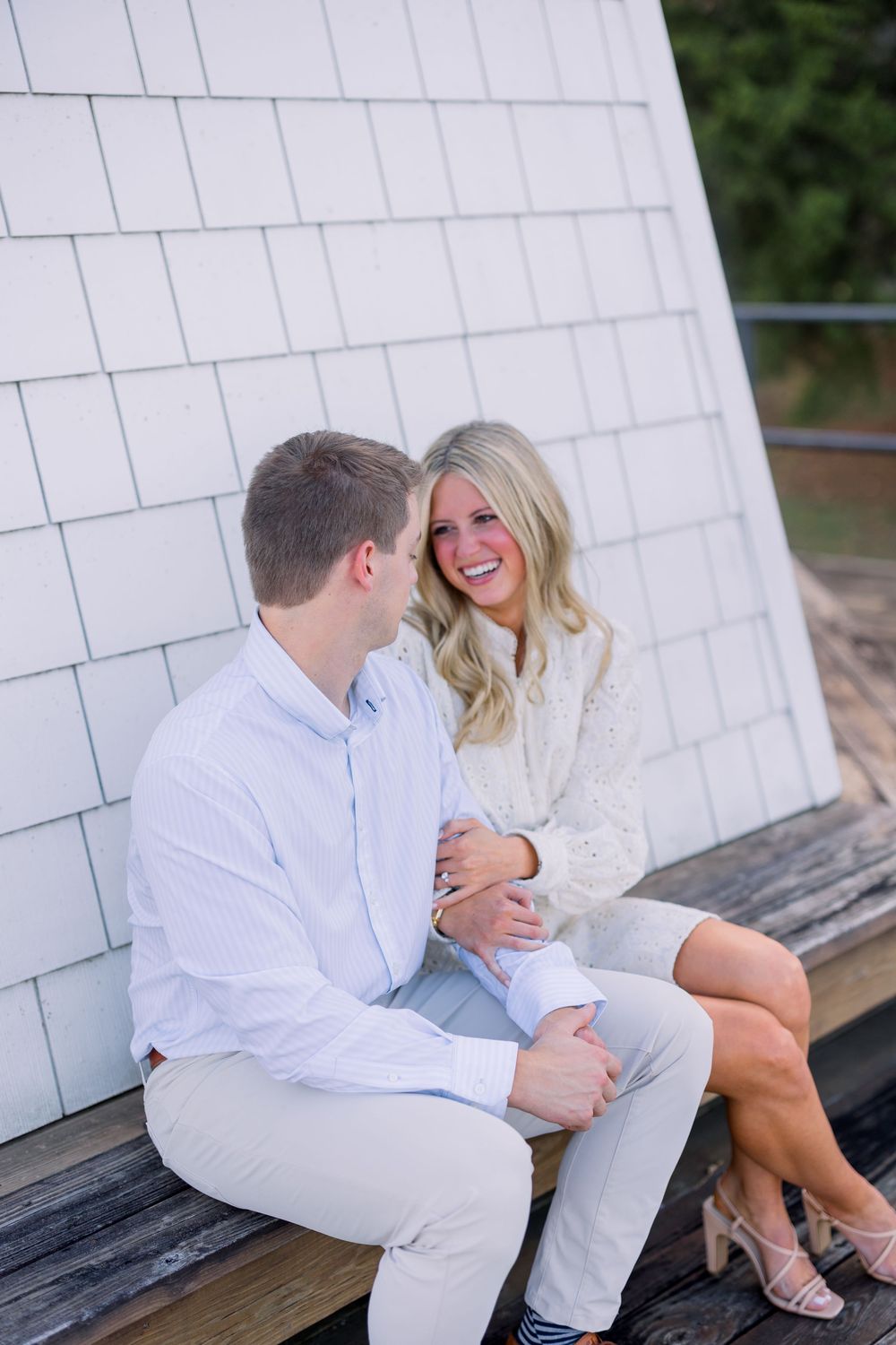 A couple wearing white outfits shares intimate moments while sitting on a wooden bench against a white wall.