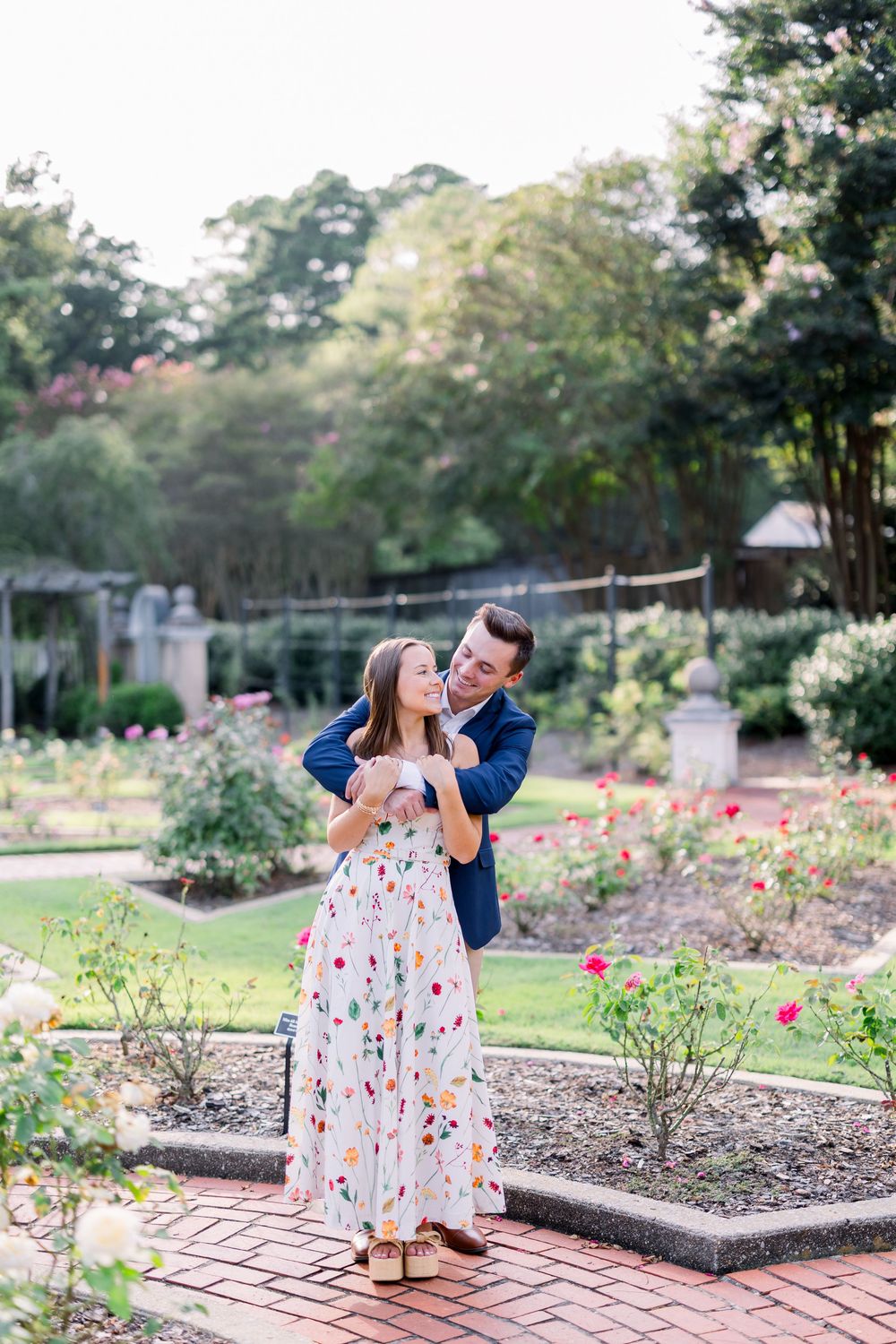 A couple shares tender moments in a blooming rose garden with raised brick planters on a sunny afternoon.