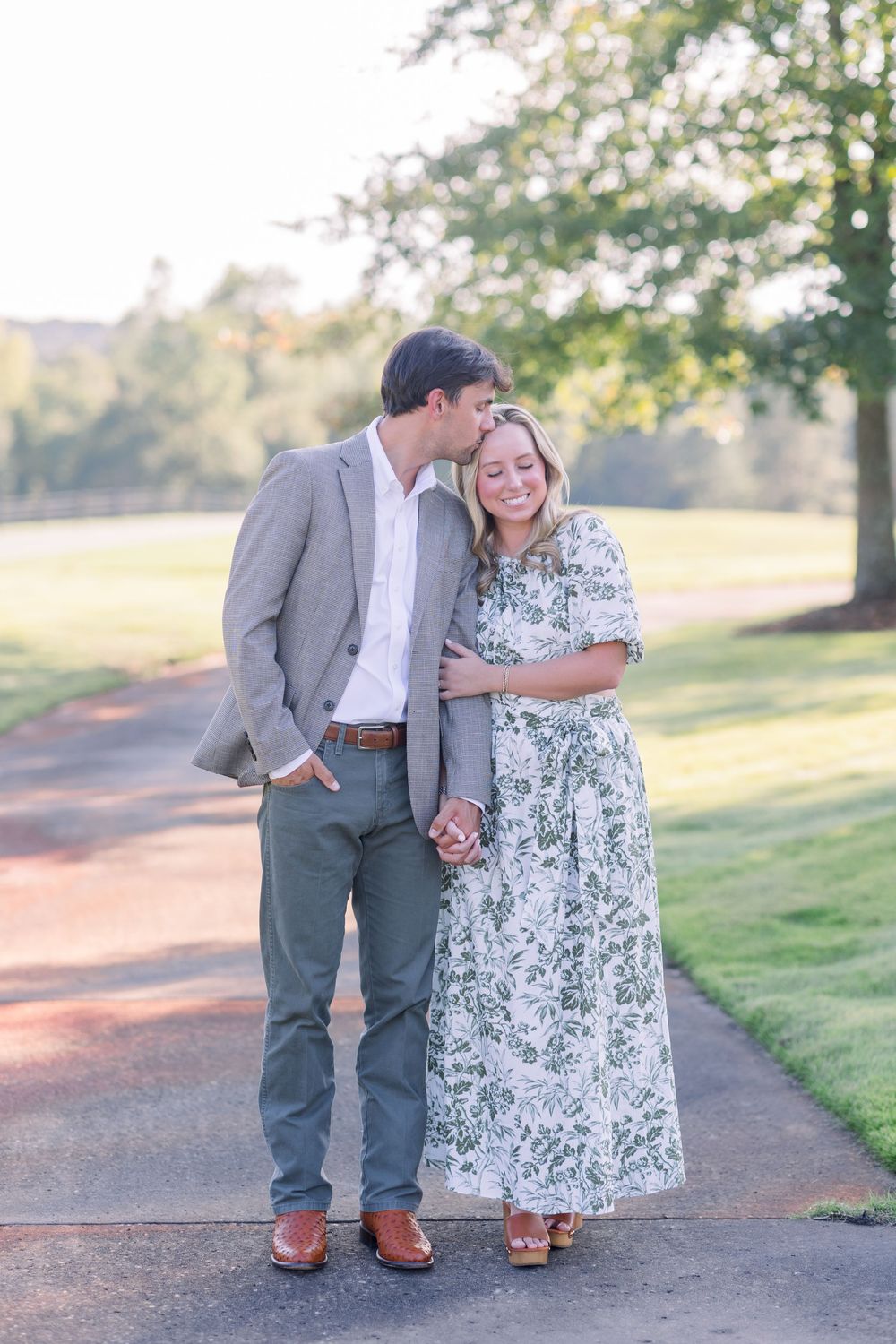 A couple in a floral dress and grey suit share romantic moments on a tree-lined path during an outdoor photo session.