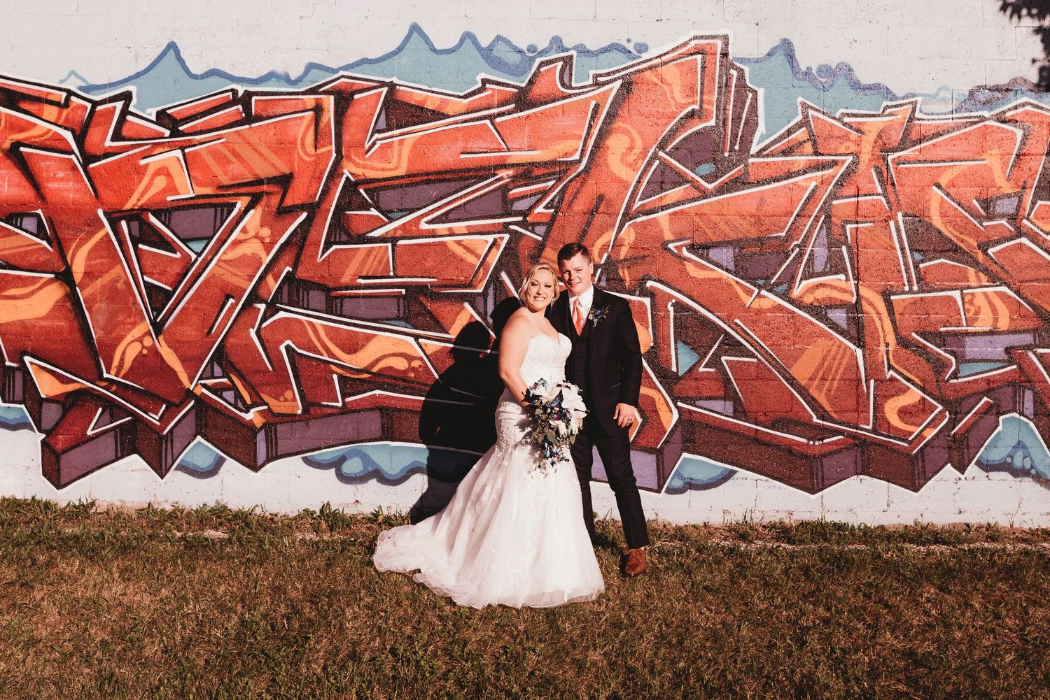 A couple in wedding attire stands against a vibrant red graffiti wall outdoors.