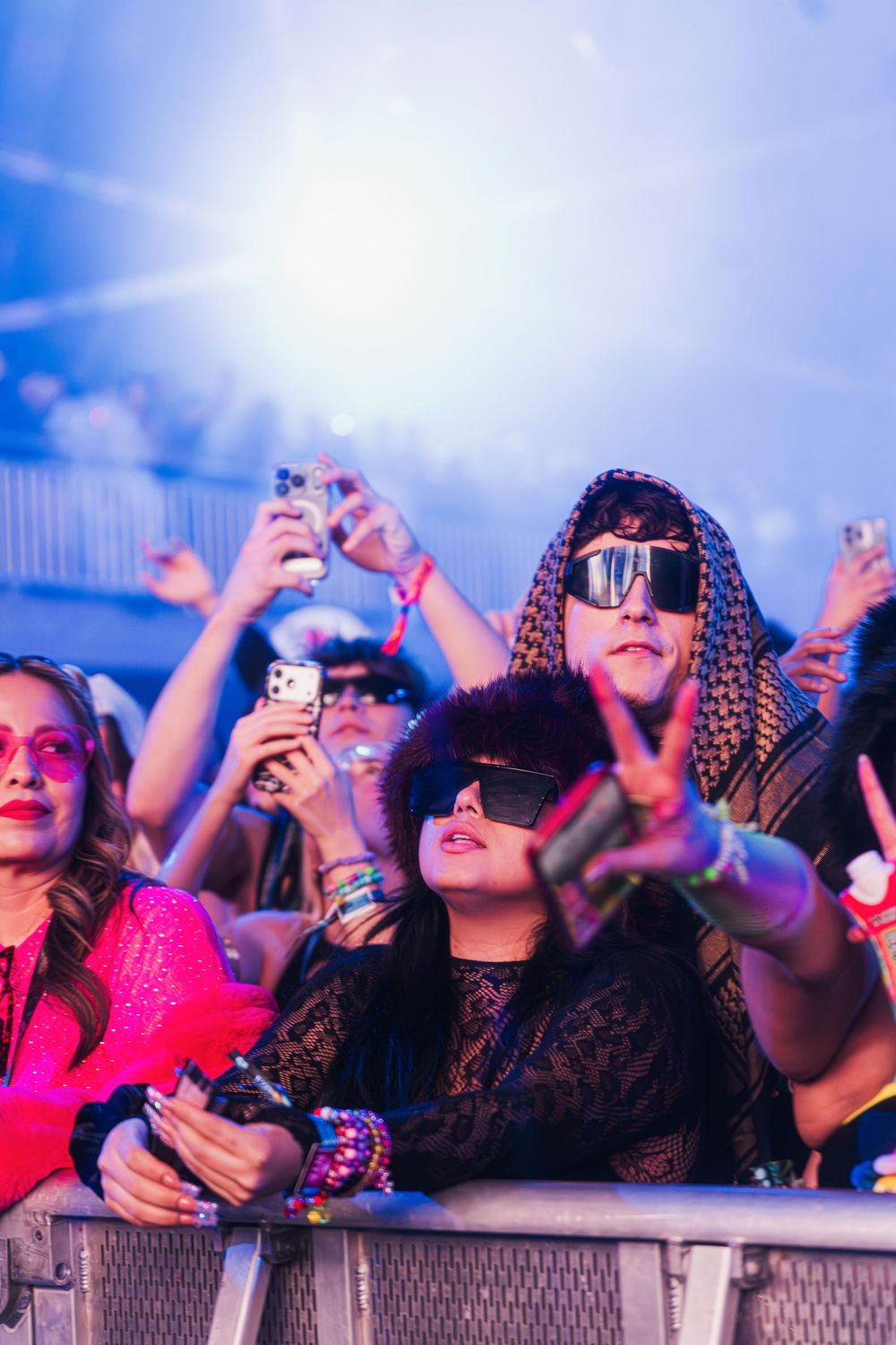 Concert crowd enjoying music festival with phones raised and sunglasses under blue sky lighting.