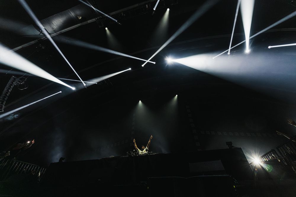 Dramatic stage lighting creates stark beams and shadows against a dark ceiling in a theater or performance space.
