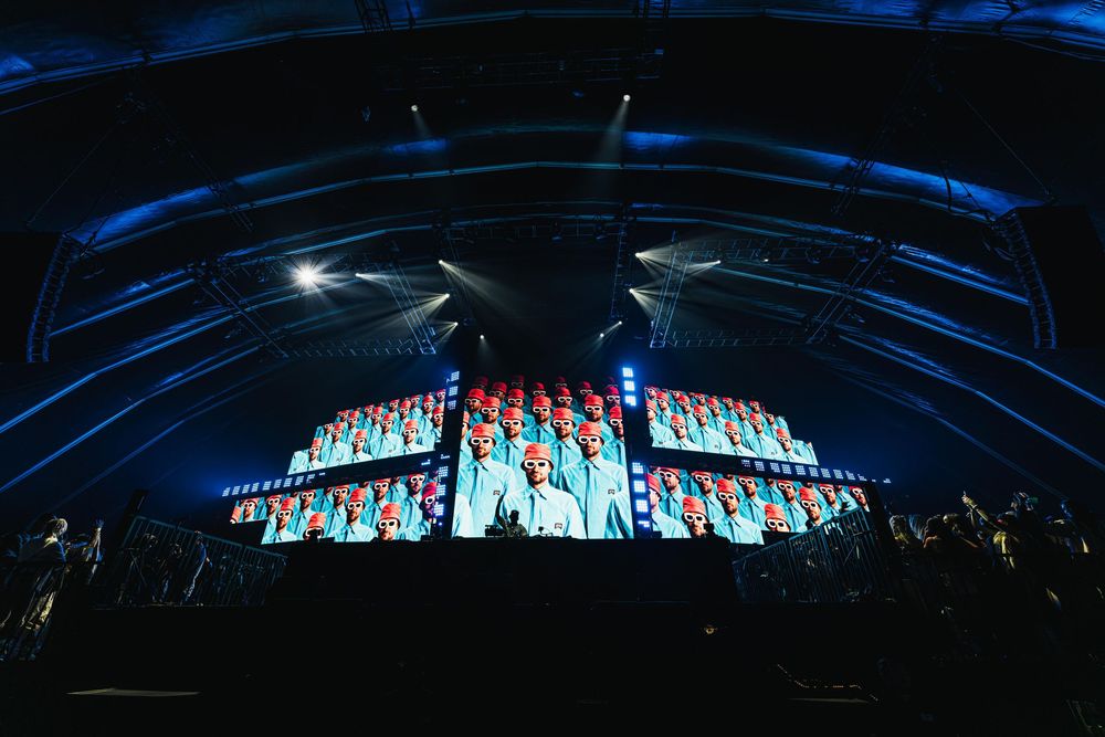 A panoramic view of a large concert stage with blue lighting and visual effects illuminating the dark venue.