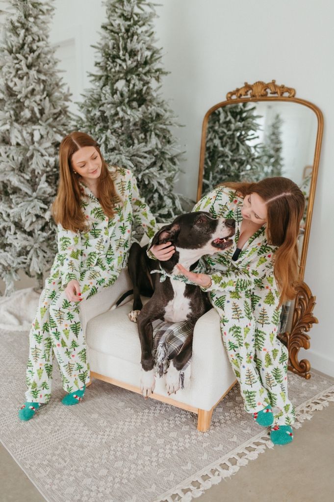 Holiday photo with matching green pine tree pajamas and a playful black and white dog on white chair by snowy trees.