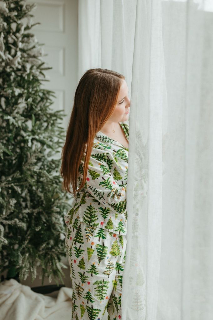 Person in festive botanical print pajamas stands by white curtains and greenery in a bright room.