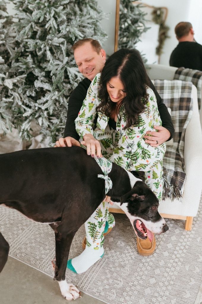 A person in a floral top plays with a large black dog on a patterned rug near a Christmas tree.