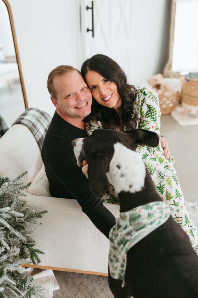 A couple sits on a couch with their pets including a black dog wearing a festive bandana and a cat during holiday season.