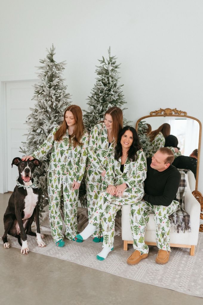 Family in matching green and white cactus print pajamas pose with their black and white dog by Christmas trees.