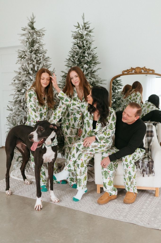 Family in matching Christmas pajamas poses playfully with their Great Dane dog in front of decorated trees.