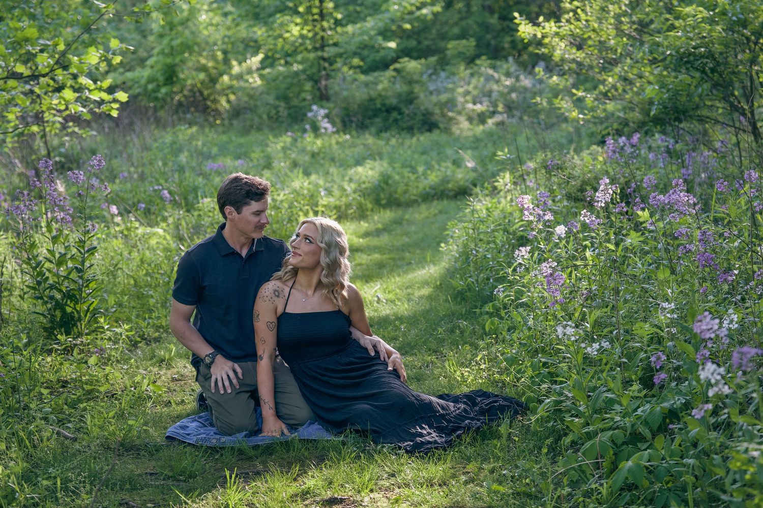 A couple sits together on a grassy path surrounded by blooming purple wildflowers on a summer afternoon.