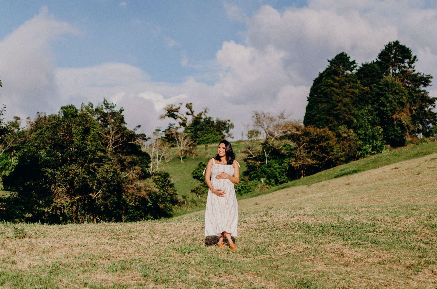Person in white dress standing on grassy hillside with trees and blue sky.