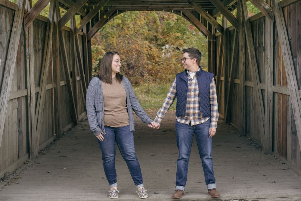 A couple holds hands while standing inside a rustic covered bridge during an autumn day.