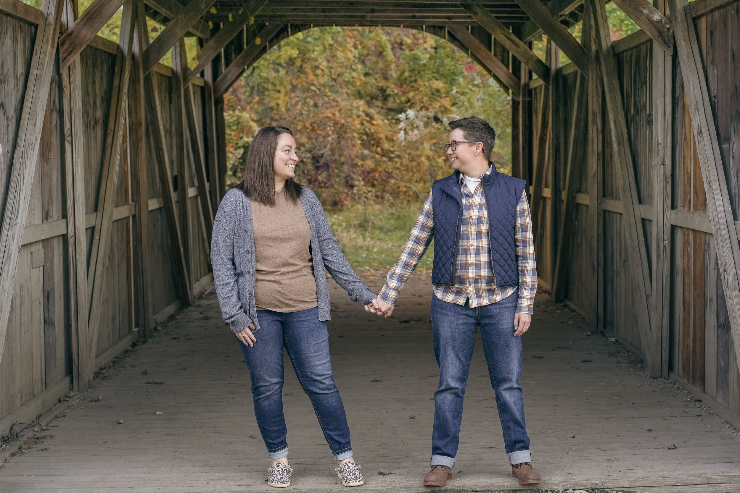 A couple holds hands while standing inside a rustic covered bridge during an autumn day.