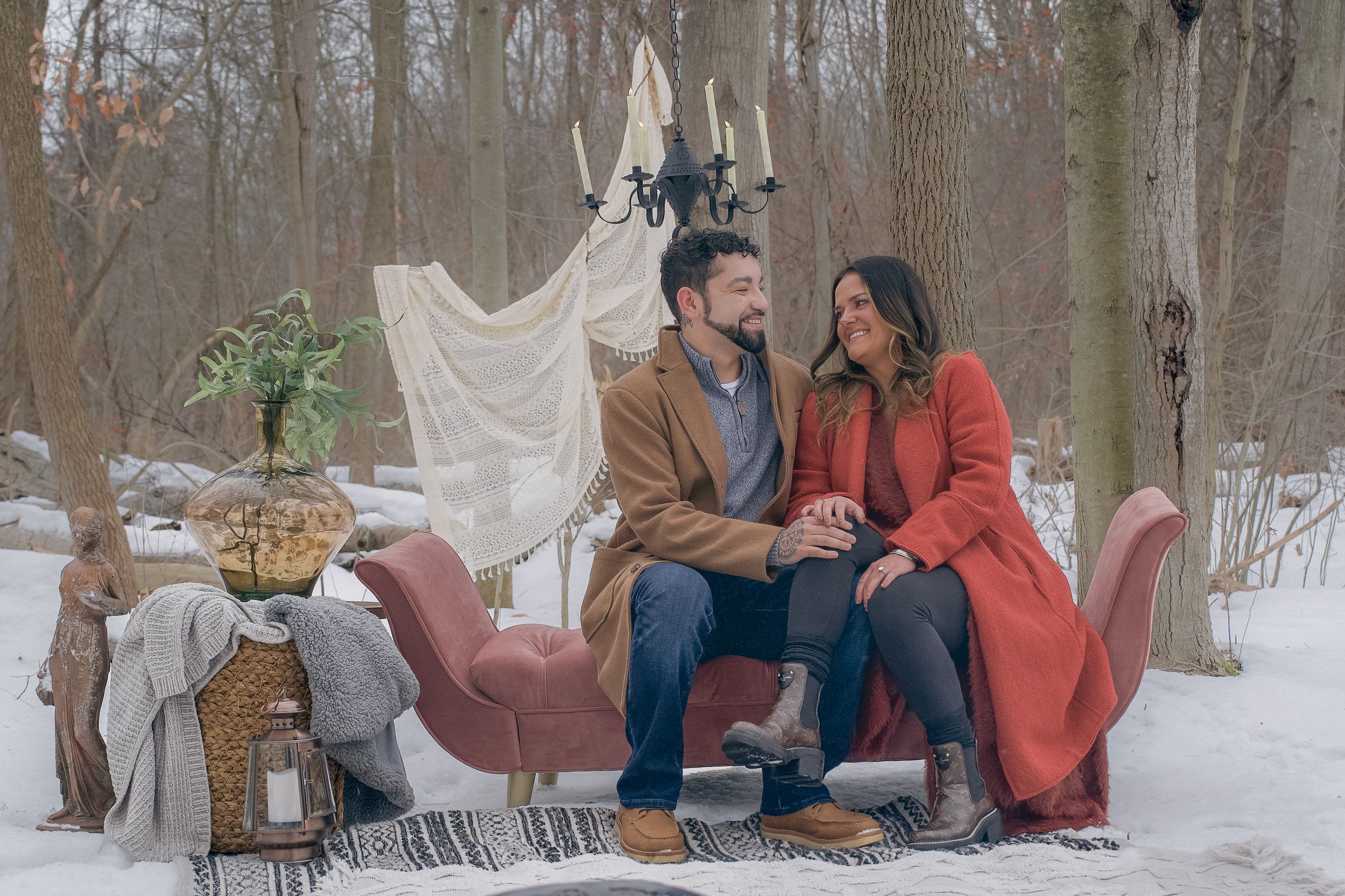 A couple sits together on a blanket in a snowy forest during a romantic winter photoshoot.