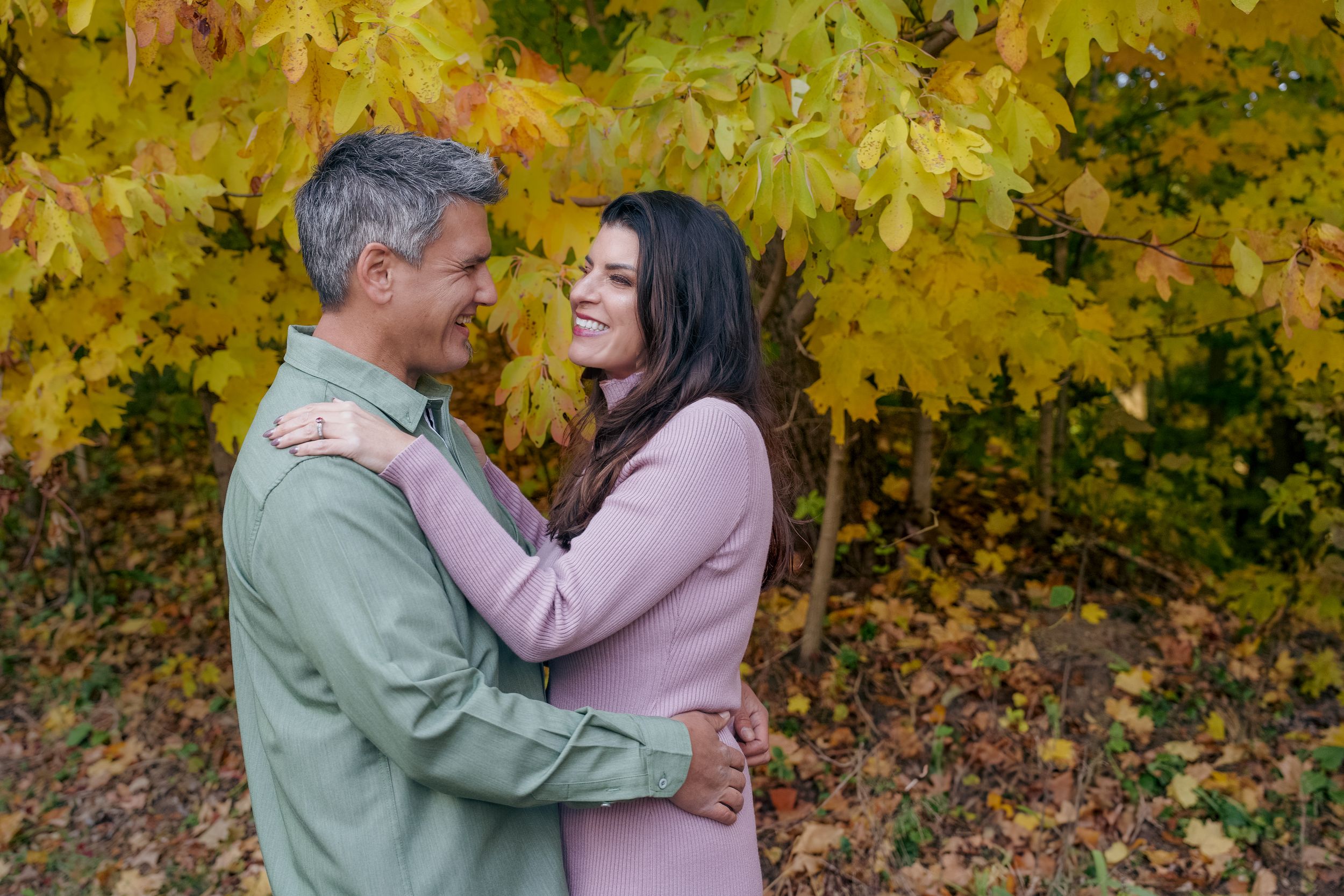 A couple embraces tenderly in front of vibrant yellow autumn foliage in a romantic outdoor setting.
