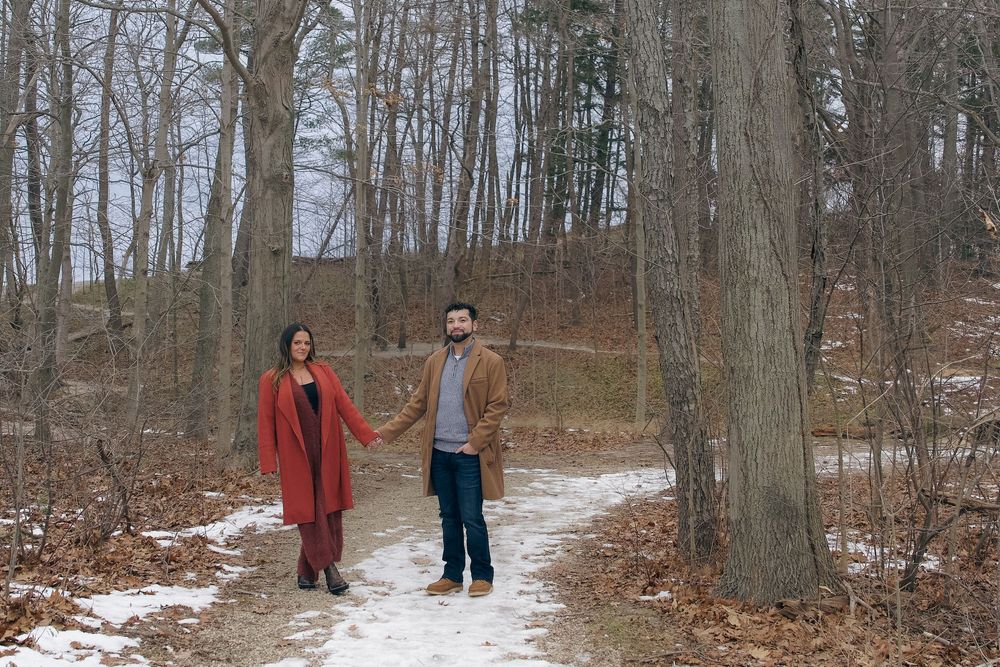 A snowy winter forest trail with two people walking through bare trees and patches of snow on the ground.