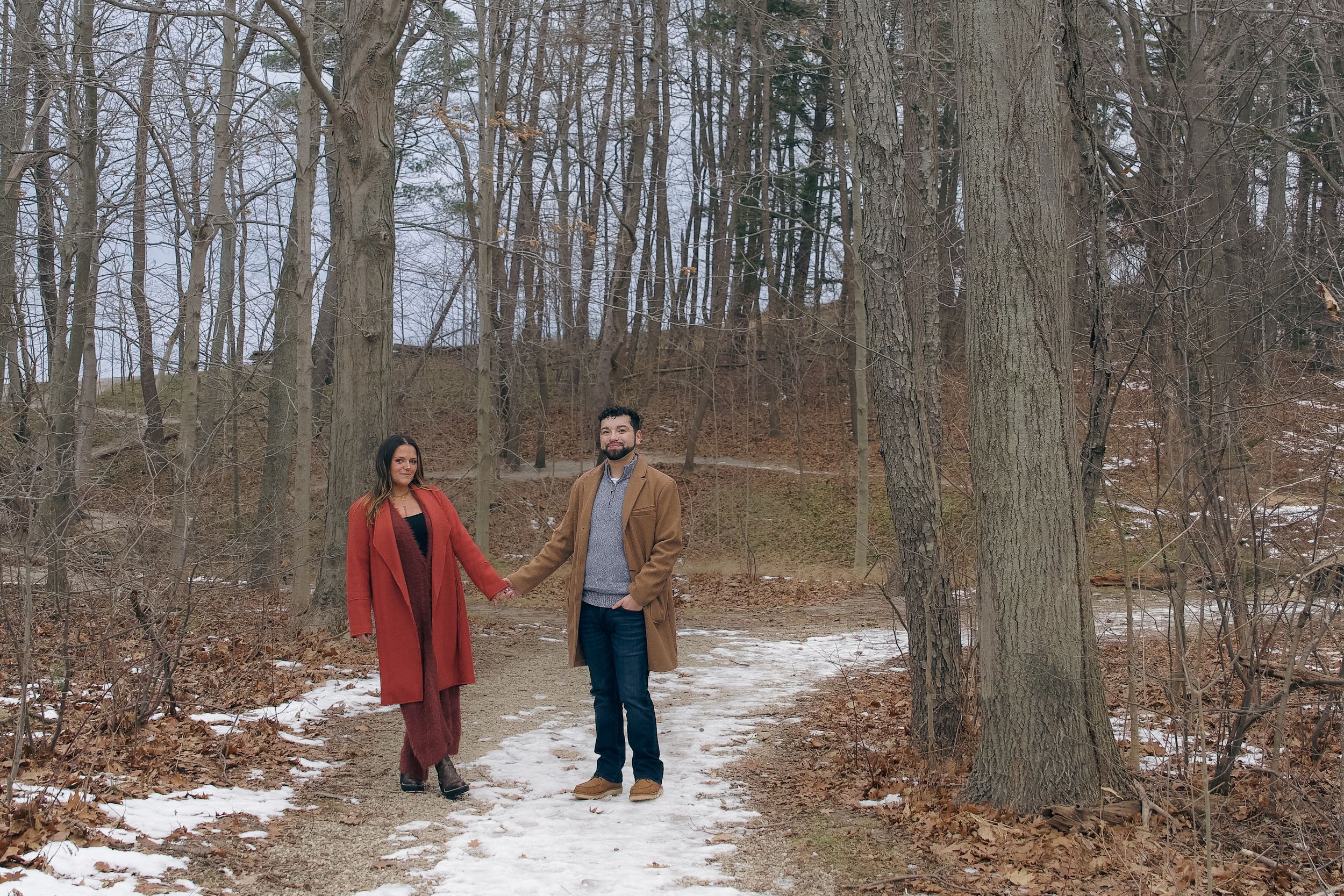 A snowy winter forest trail with two people walking through bare trees and patches of snow on the ground.