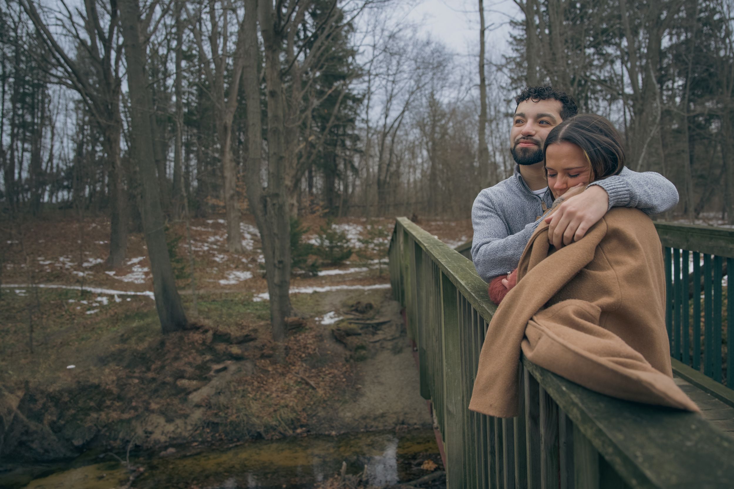 Two people share a tender moment together while sitting outdoors on wooden steps surrounded by trees and foliage.