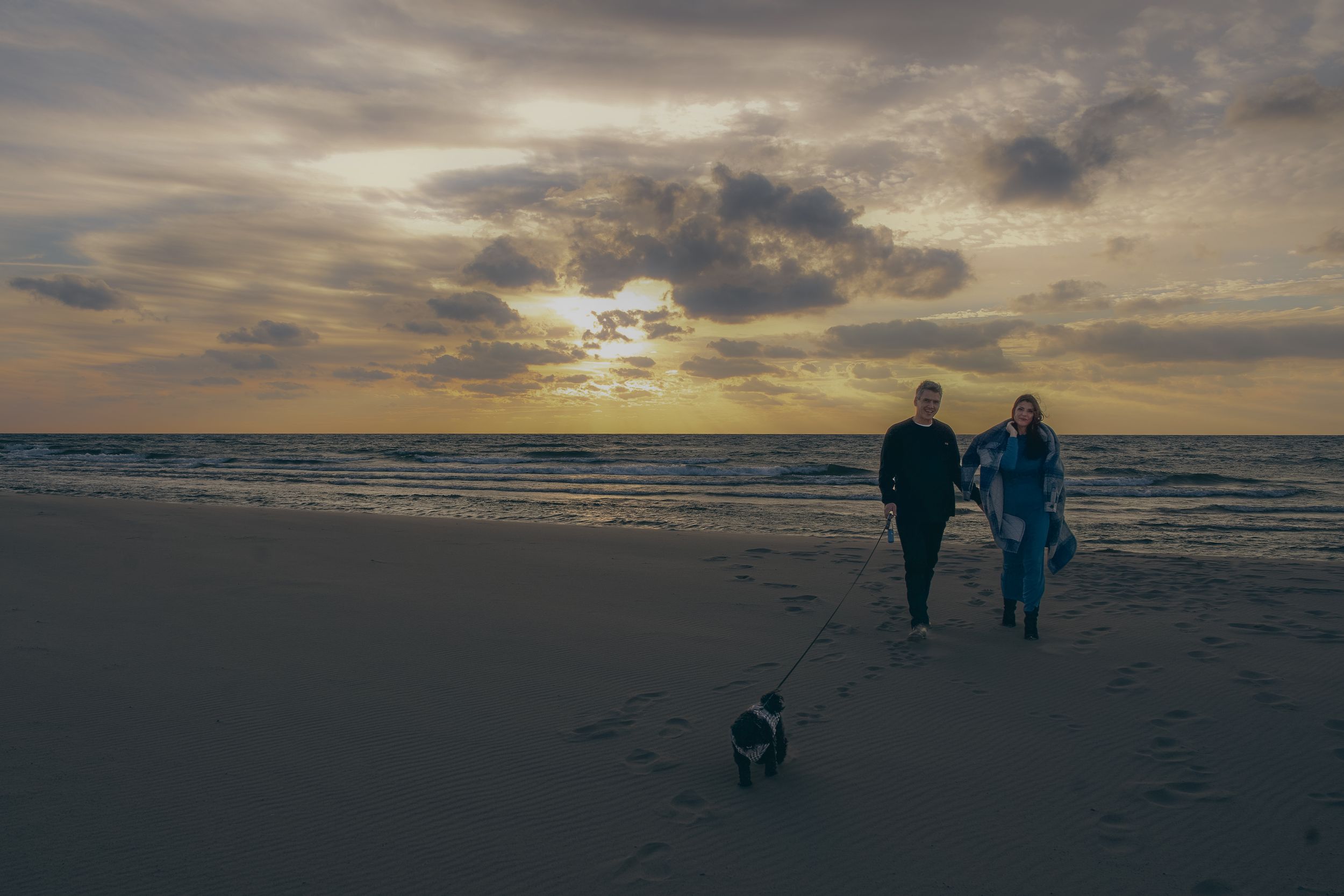 Silhouettes of two people walking their small dog on a beach during a dramatic cloudy sunset.