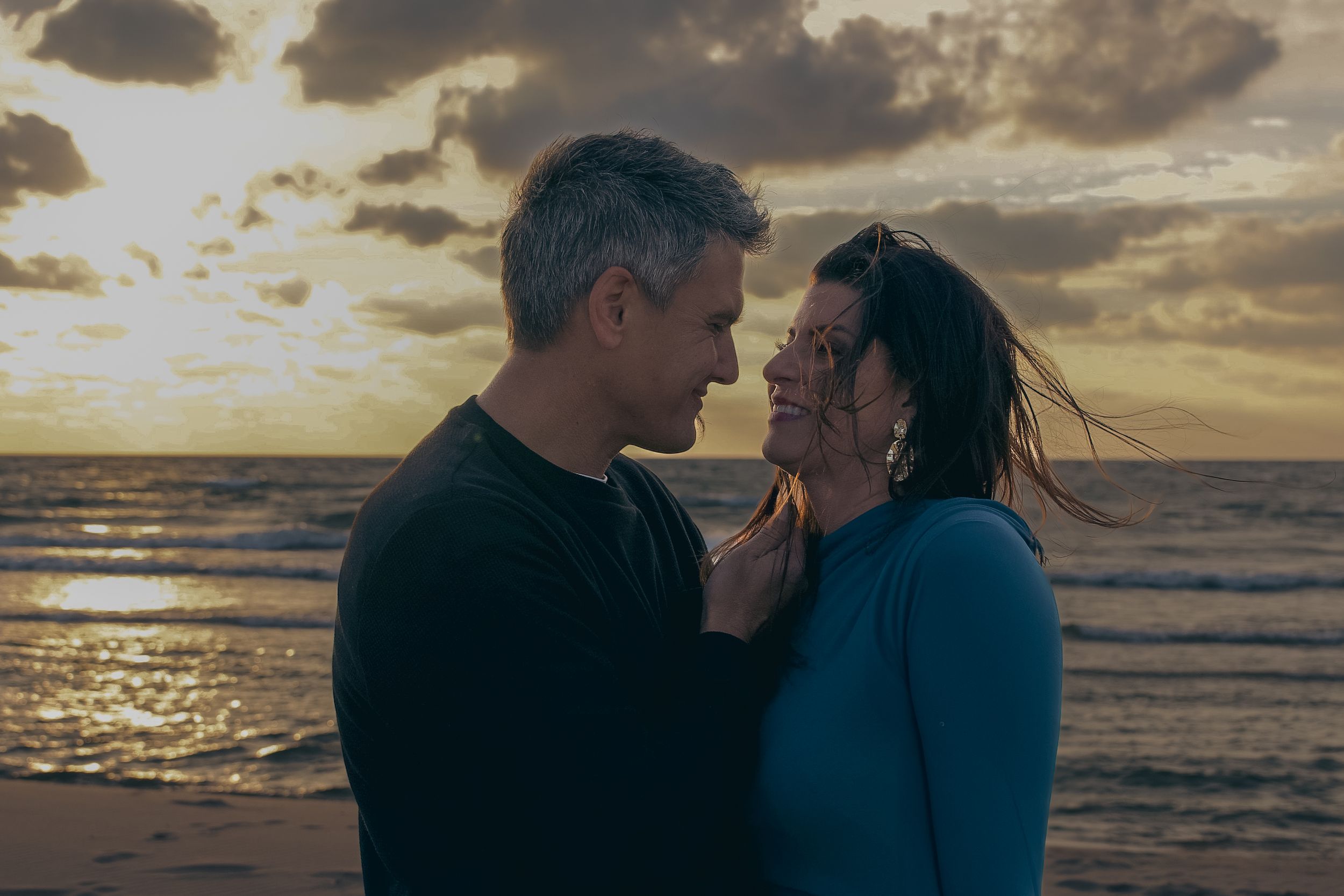 A romantic couple embraces on a beach at sunset with dramatic golden clouds in the sky.