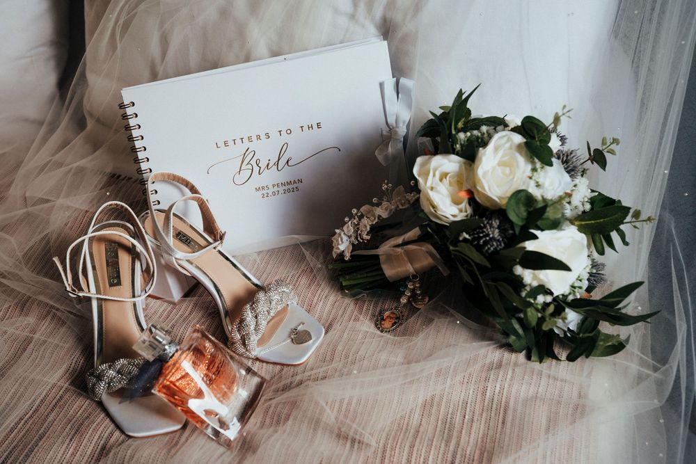 Wedding details flatlay with white floral bouquet, strappy heels, perfume bottle and vow book on blush fabric.