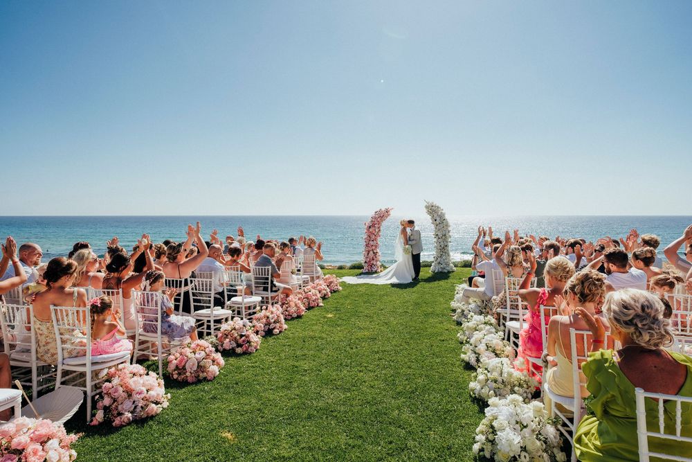 Wedding guests seated in white chairs on a grassy hillside overlooking a scenic mountain landscape during an outdoor ceremony.