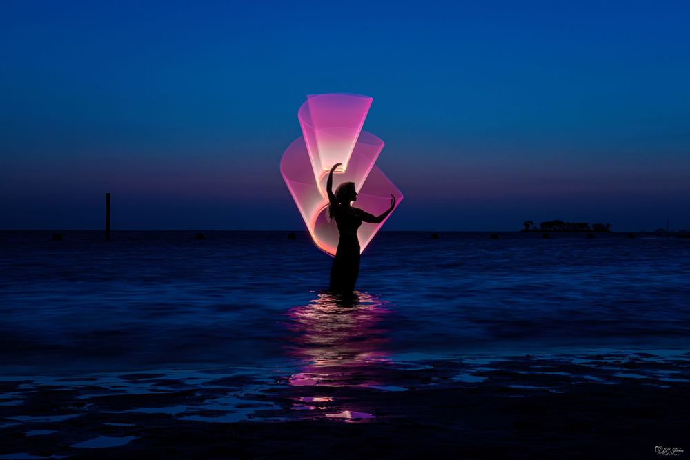 Silhouetted figure creates luminous patterns against a deep blue coastal dusk landscape.