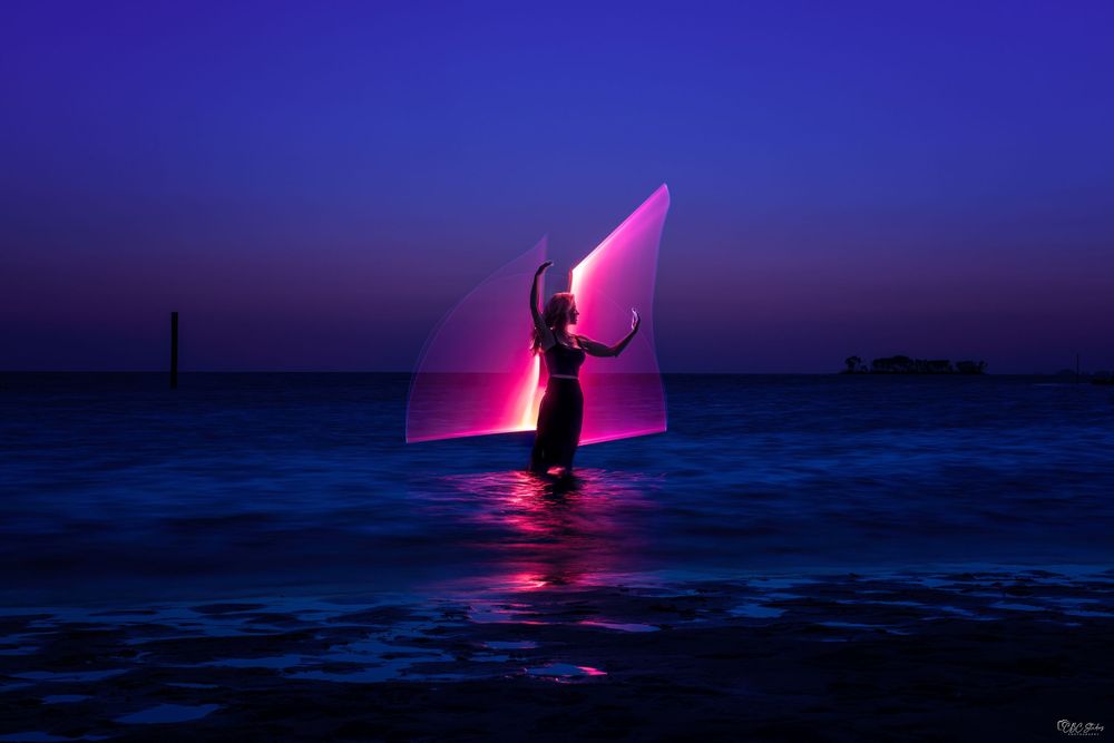 Silhouetted figure dances on a beach against vibrant purple sunset reflecting on calm ocean waters.