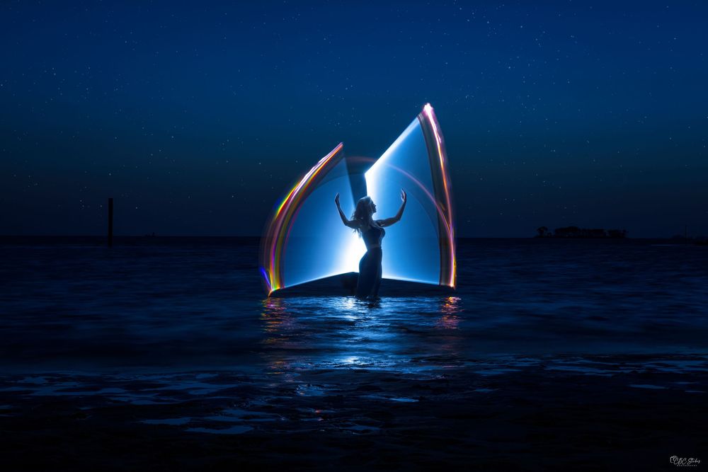 Illuminated sailboat glides across dark waters under moonlight creating a mystical reflection on the ocean surface.