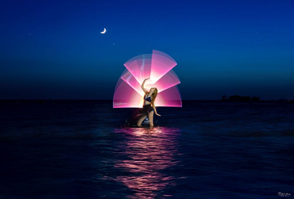 A couple embraces on a boat at sunset while a pink sun creates a dramatic silhouette against the ocean horizon.