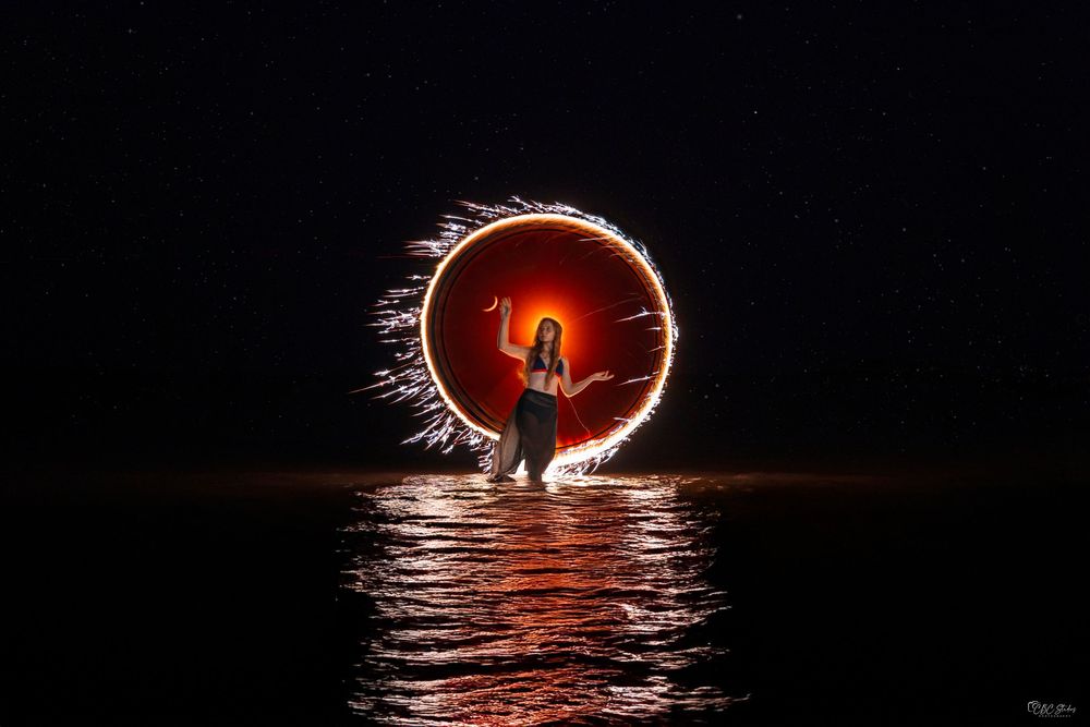 Long exposure photograph shows light painting creating a circular pattern over dark water at night.
