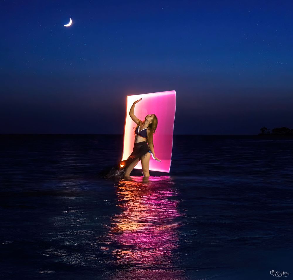 A glowing pink neon light frame illuminates the dark ocean at night, creating a surreal and artistic scene.