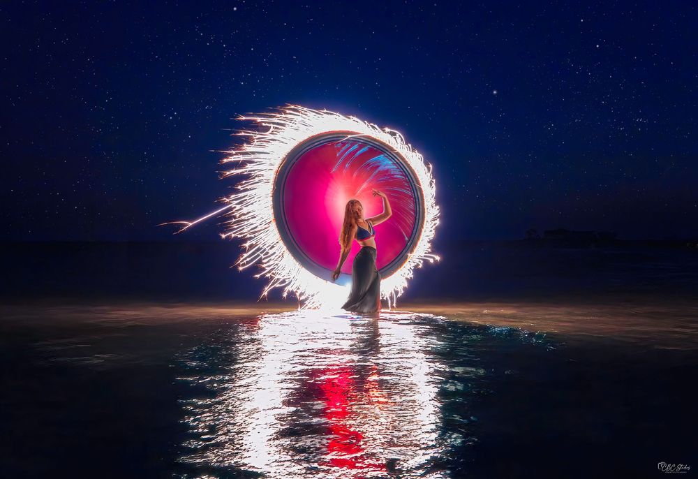 Long exposure photography captures spinning steel wool creating circular light trails reflected on water at night.