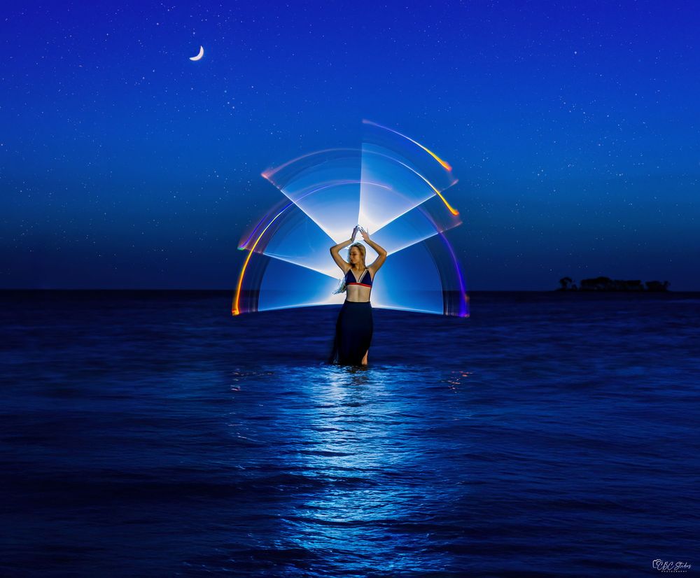 Steel wool light painting creates luminous arcs over calm water against a deep blue twilight sky.