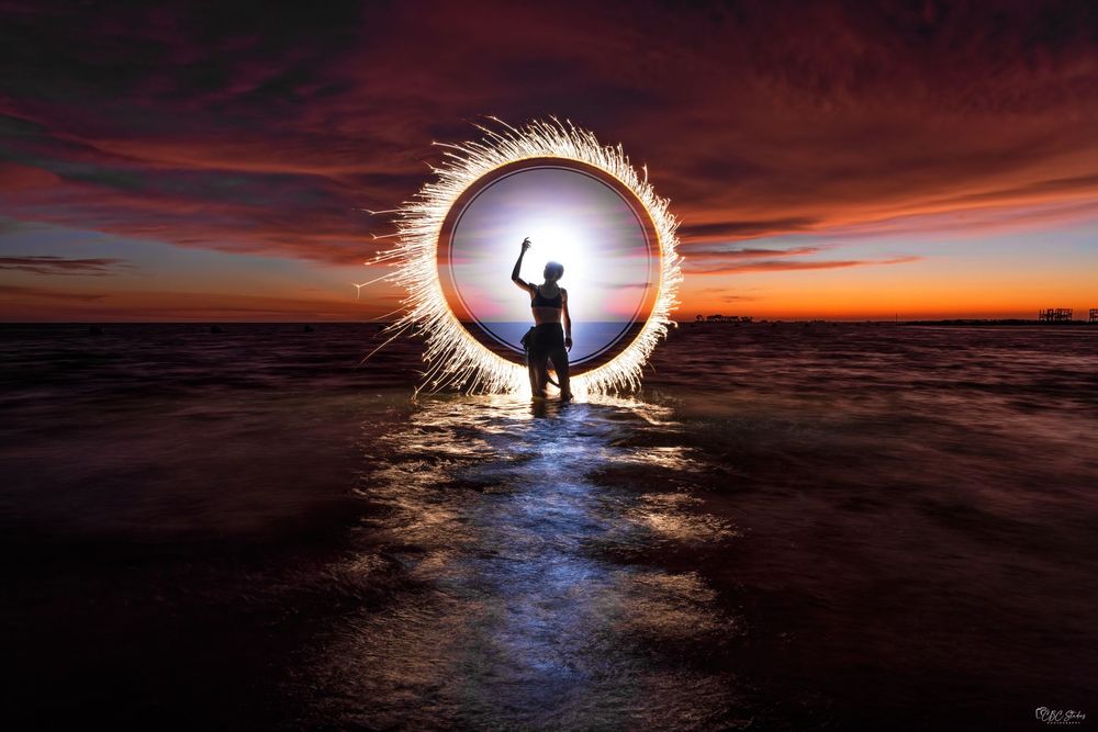 A silhouetted figure stands in a glowing light circle on water against a dramatic sunset sky.