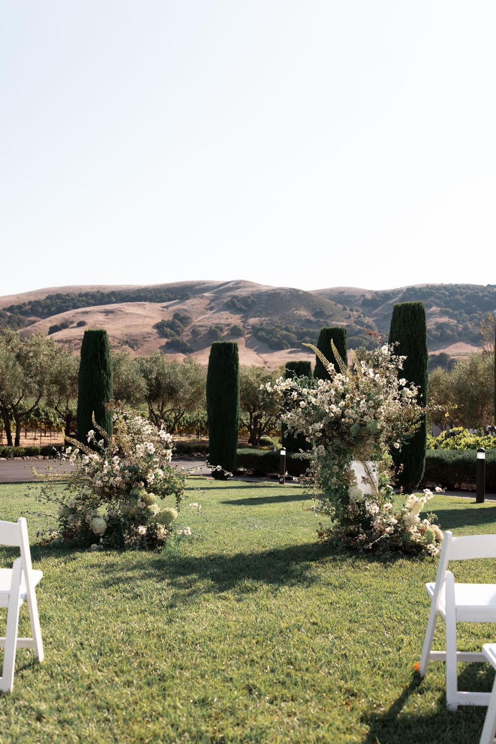 White chairs arranged on grass lawn with flowering trees and mountains in the distance.