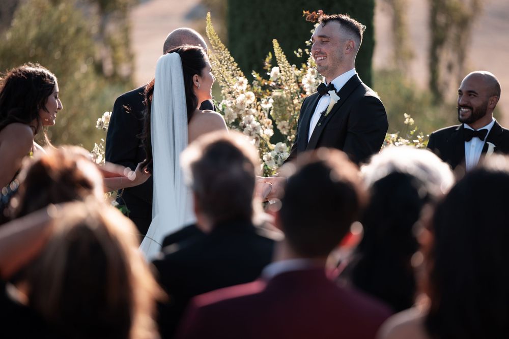 A wedding ceremony takes place outdoors with guests gathered around as sunlight filters through trees in the background.