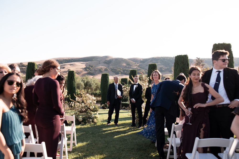 Wedding guests gather at an outdoor ceremony venue with rolling hills and scenic views in the background.