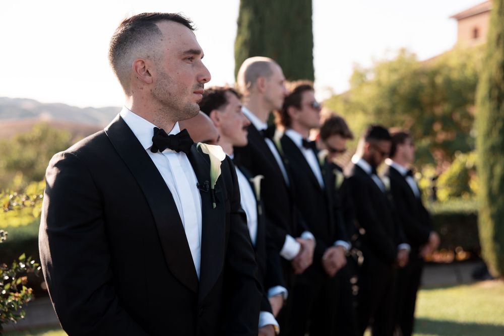 A row of groomsmen in black tuxedos stand outdoors during a wedding ceremony at sunset.