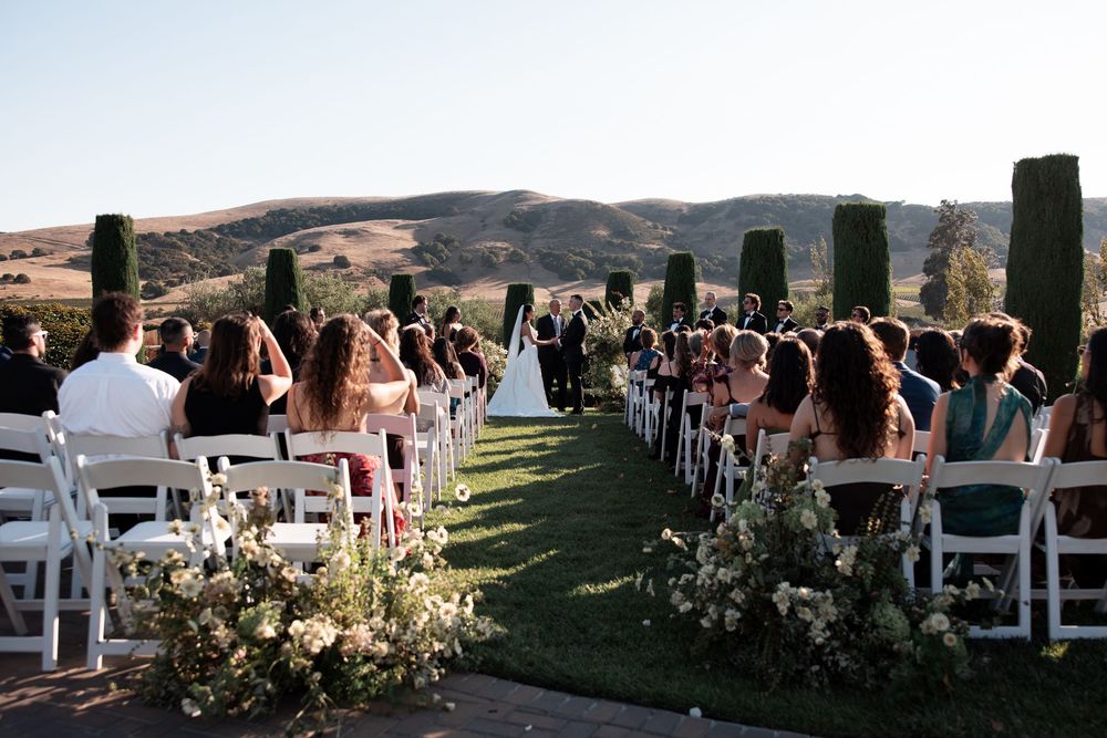 Outdoor wedding ceremony with white chairs and floral arrangements set against rolling hills and stone pillars.