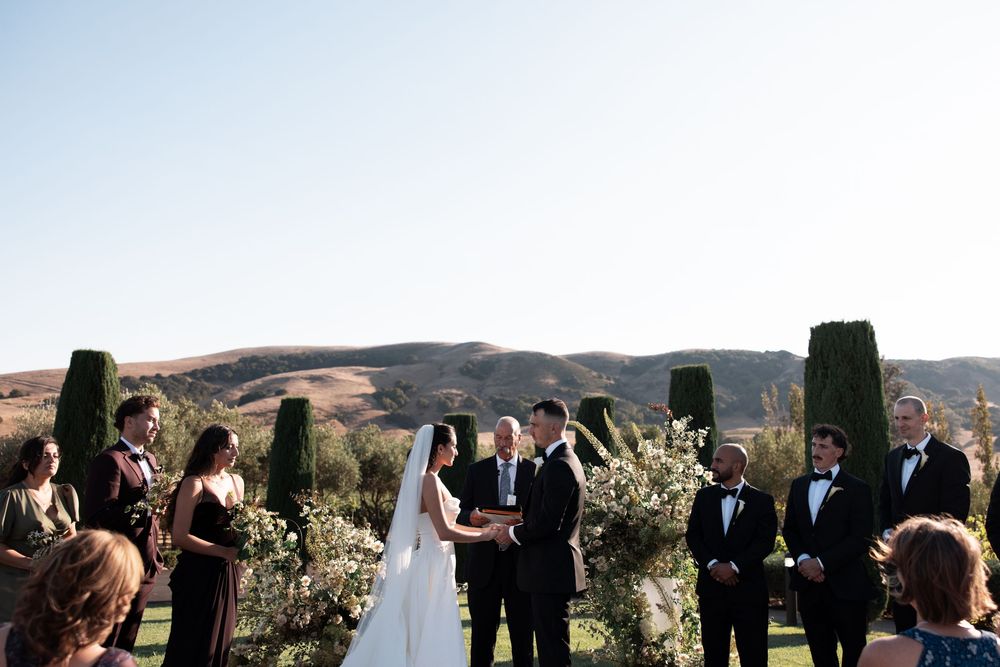 Wedding ceremony takes place outdoors with guests gathered on a scenic hilltop overlooking a mountain landscape.