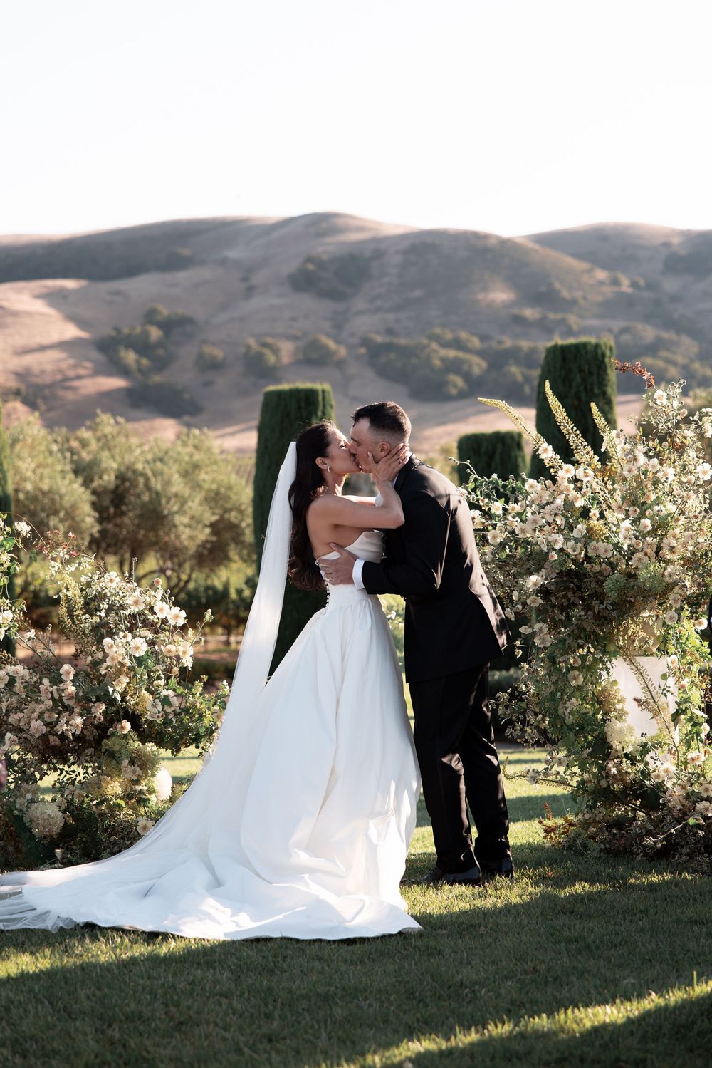 A romantic wedding couple shares tender moments in a garden setting with rolling hills and greenery in the background.