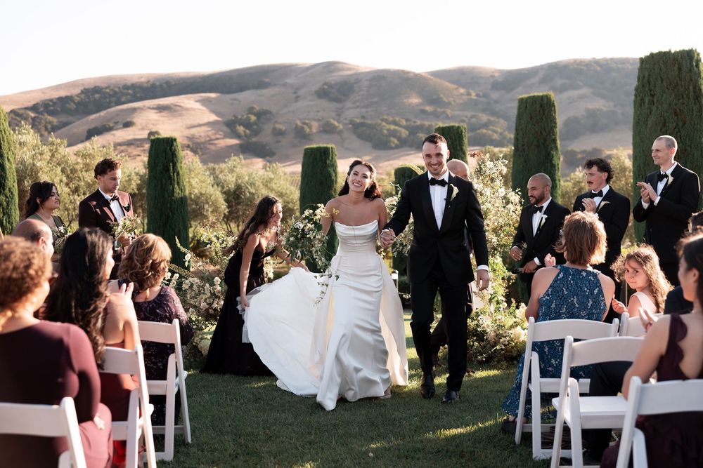 Panoramic view of an outdoor wedding ceremony with white chairs set up on a hillside overlooking mountain vistas.