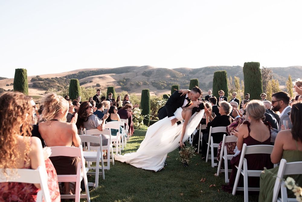 Outdoor wedding ceremony at sunset with guests seated in white chairs overlooking scenic mountain views.