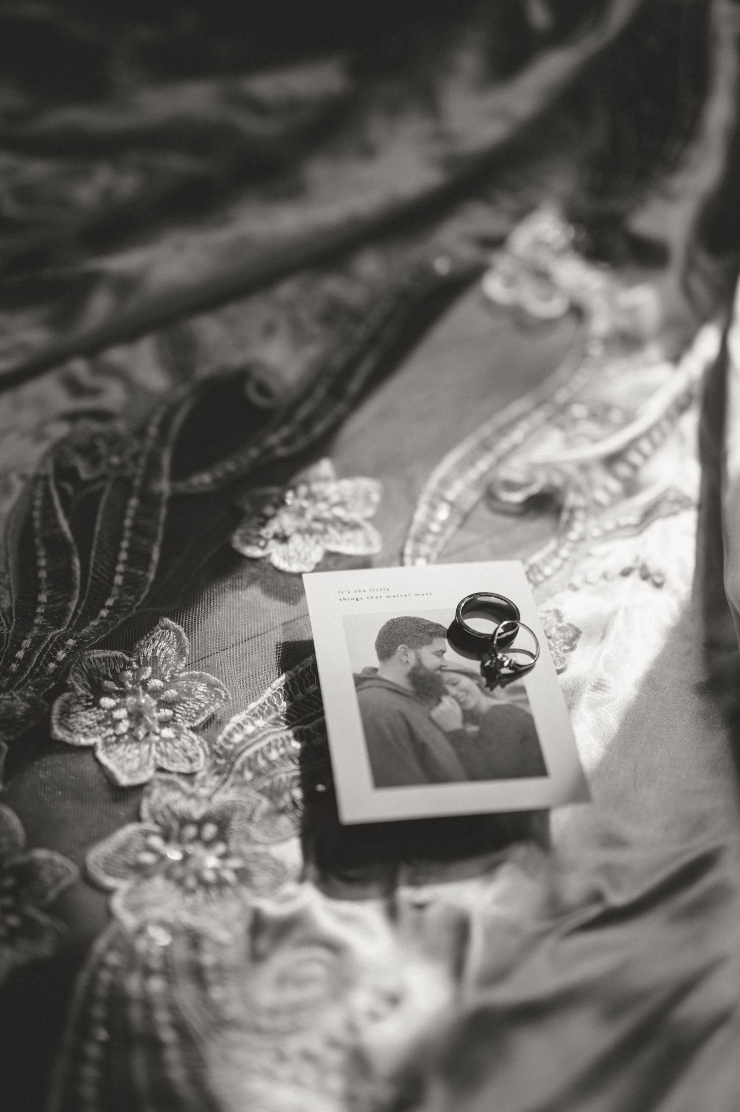 Black and white photo of wedding rings resting on a couple's portrait atop an elegant lace bridal gown.