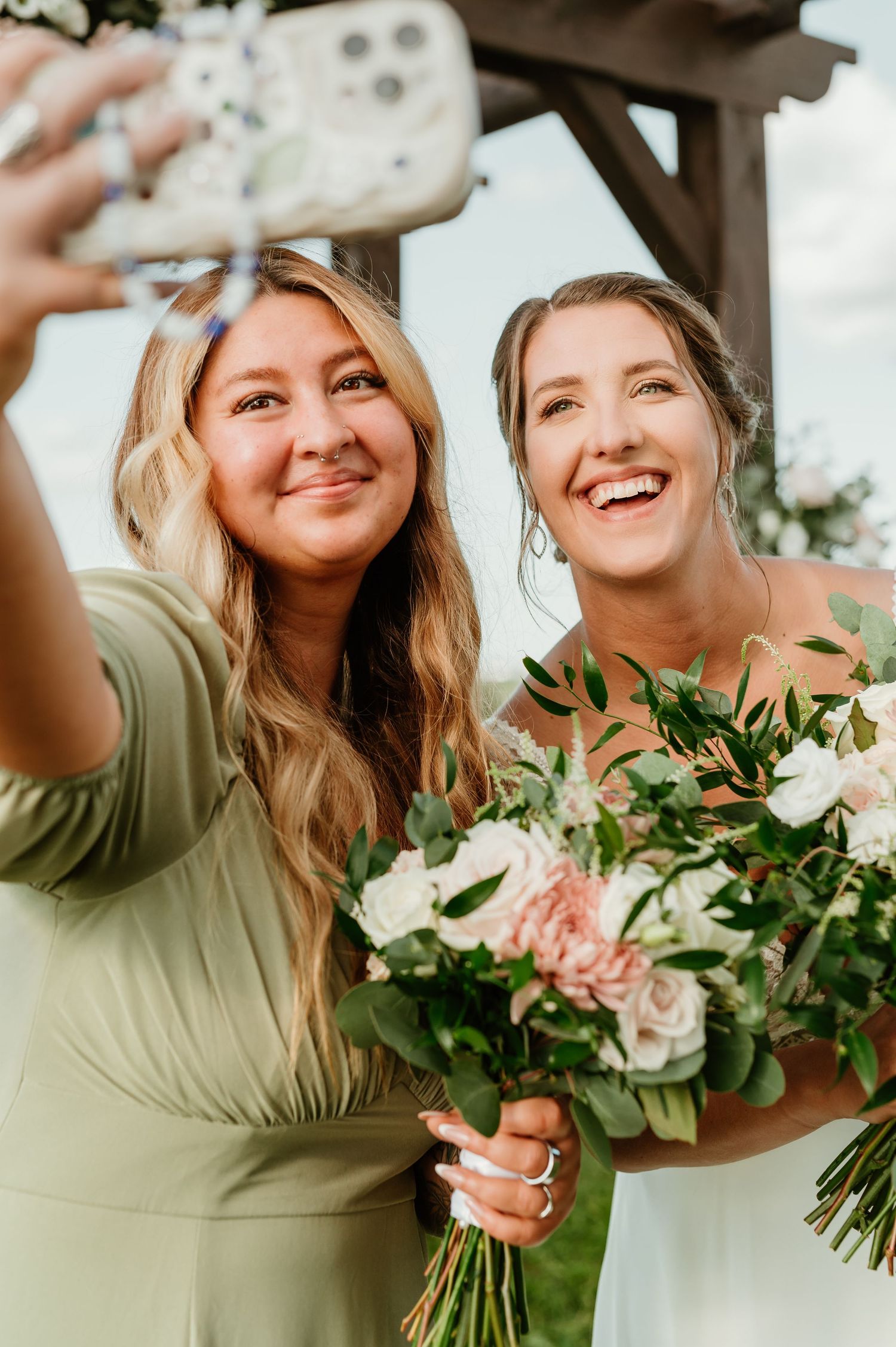 Bride and bridesmaid in sage green dress taking a selfie while holding pink and white floral bouquets outdoors.