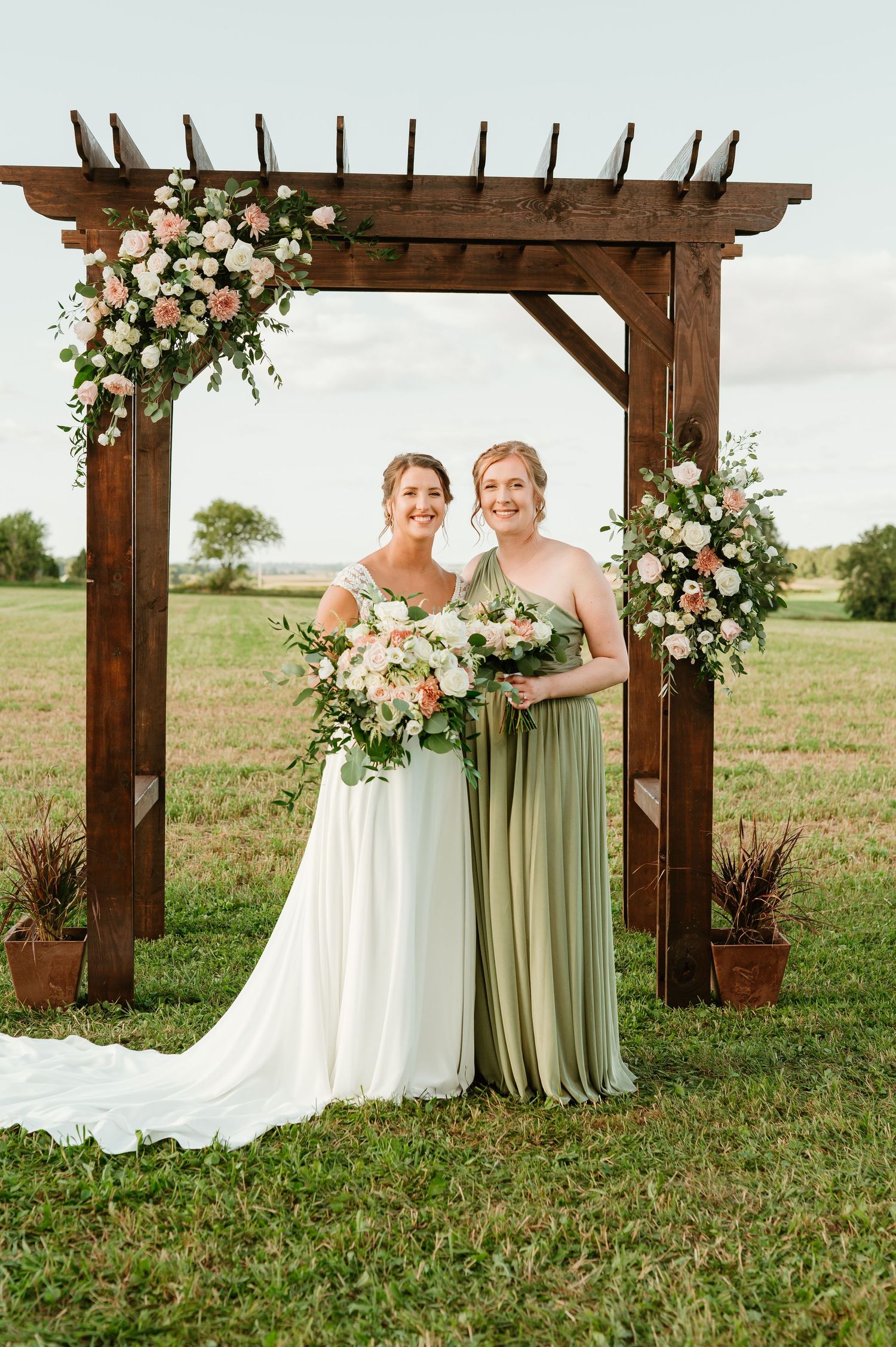 Bride in white gown and bridesmaid in sage green dress pose under rustic wooden arch with pink floral arrangements.